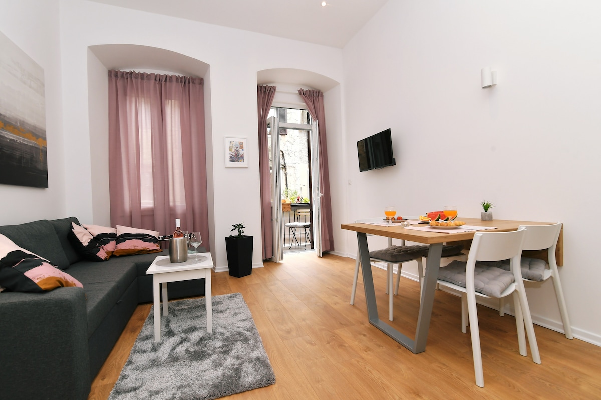 A spacious living area features a gray sofa adorned with colorful cushions. A dining table with white chairs is set for a meal, with bright drinks and snacks visible. Natural light enters through large windows dressed with light curtains, illuminating the cozy ambiance.