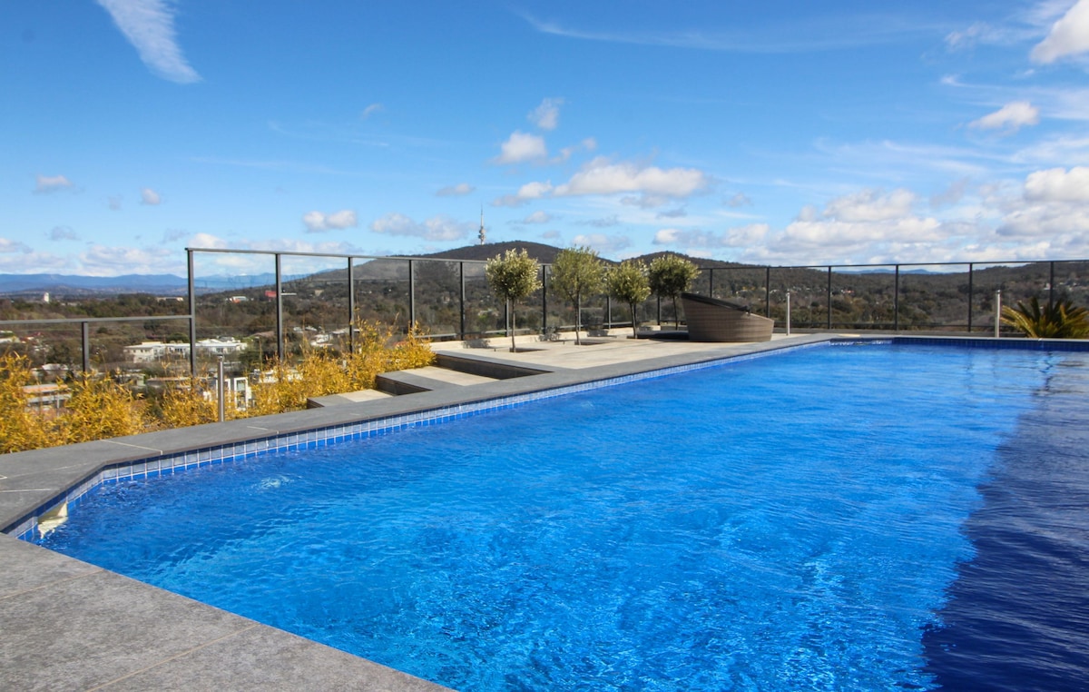 The rooftop area features a spacious salt-water pool, surrounded by a glass railing. Sunlight reflects off the water's surface, with a backdrop of rolling hills and blue skies. Planters with greenery are positioned around the pool, enhancing the relaxed atmosphere.