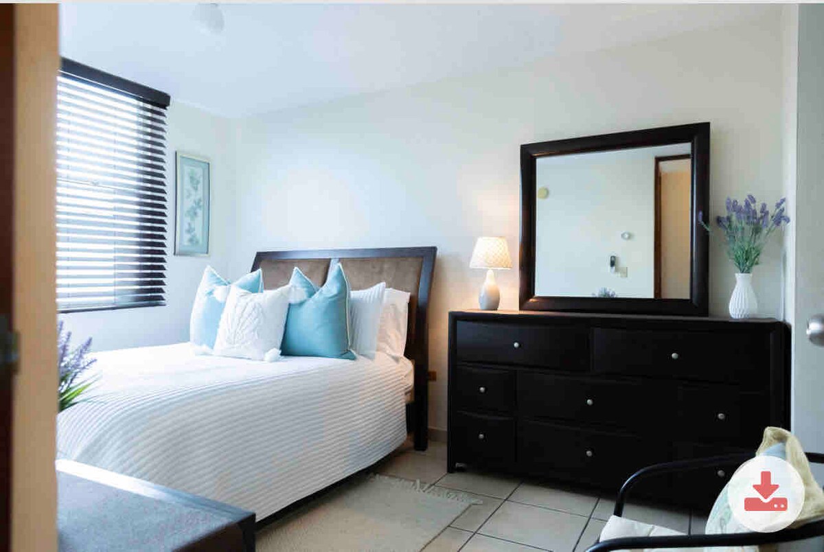 A cozy bedroom features a spacious bed adorned with decorative pillows and a light blanket. A dresser and a mirror are positioned against the wall, while natural light filters through the slatted blinds. A bedside lamp adds subtle illumination.