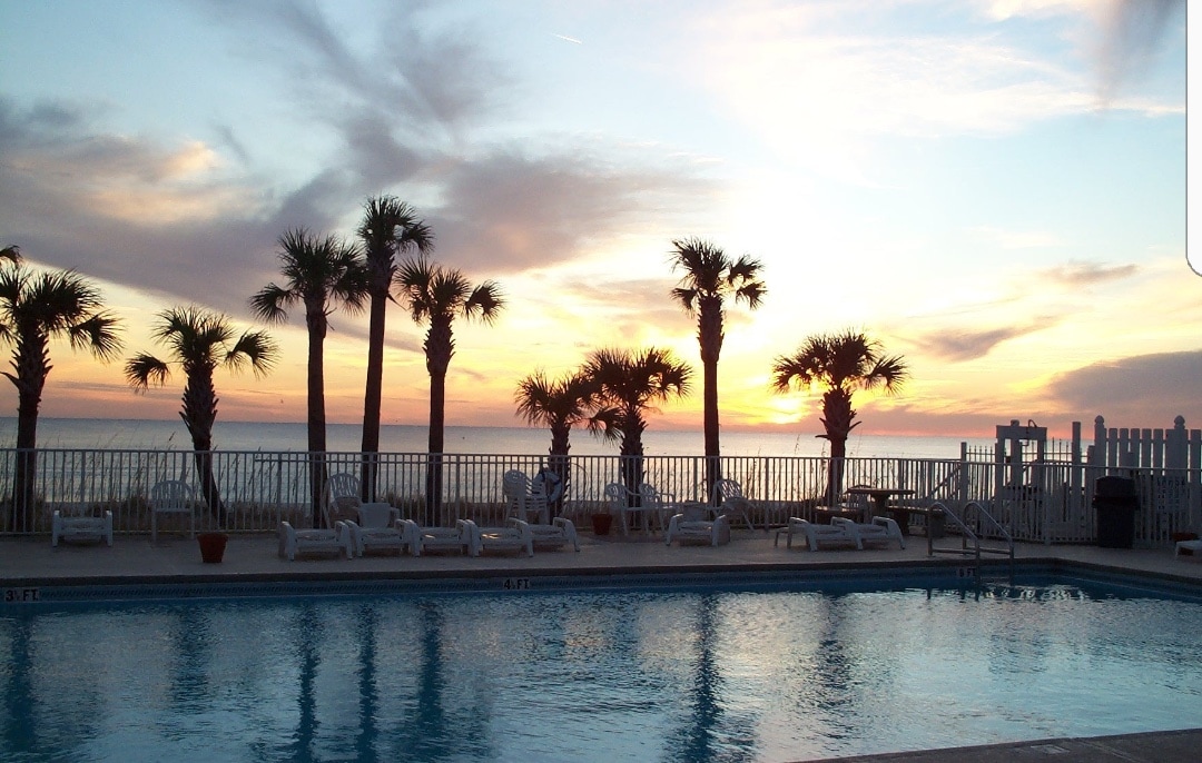 A serene pool area is visible, framed by tall palm trees against a sunset sky. Lounge chairs surround the pool, reflecting the tranquil atmosphere. The ocean is seen in the background, creating a picturesque view of the beachfront location.