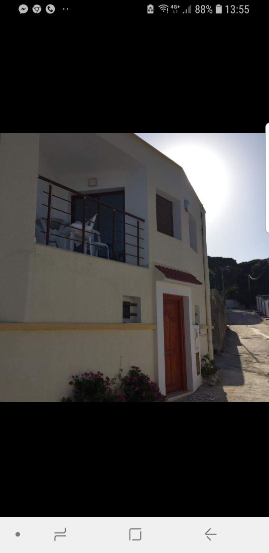 A modest structure is depicted, featuring a light-colored exterior and a welcoming entrance door. A small balcony with white chairs overlooks the area, while a flower bed with greenery adds a touch of nature to the space.