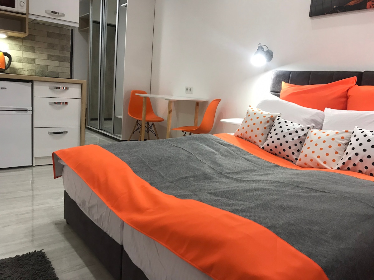 The inviting sleeping area features a spacious bed with a vibrant orange and gray coverlet. A small dining table with orange chairs is visible in the background, alongside a well-equipped kitchen area with modern appliances. Natural light fills the room from nearby windows.