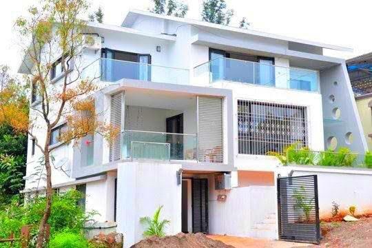 The modern villa exterior features clean lines and large glass windows, allowing natural light to fill the interior. A spacious terrace is visible, complemented by greenery and elegant landscaping. A gated entry adds privacy and enhances the overall appeal of the property.