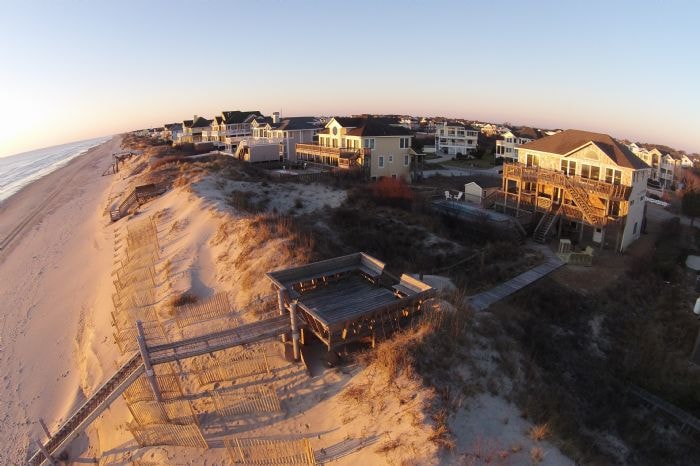 An aerial view captures the surrounding beach area, highlighting golden sand and gentle ocean waves. Nearby homes are visible, framed by natural dunes, with a wooden deck leading toward the beach. The sunset casts a warm glow over the serene landscape, enhancing the coastal charm.