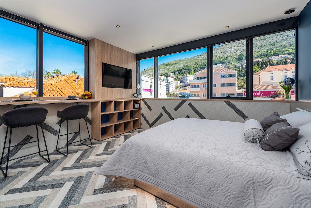 A modern studio apartment features a spacious bed against a backdrop of large windows that allow natural light to fill the room. Stylish seating at a small bar area complements the decor, while views of the surrounding city and mountains are visible through the windows.