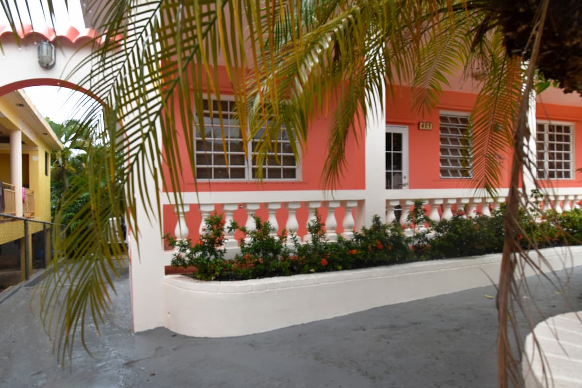 A vibrant exterior view of the house features coral pink walls and decorative white railings. Lush greenery, including palm fronds and flowering plants, frames the entrance, creating a refreshing visual contrast against the colorful facade.