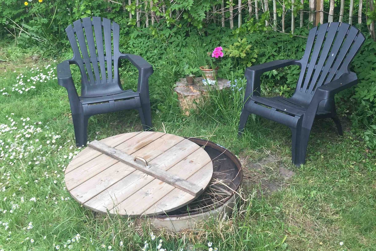 Two black Adirondack chairs are positioned on a grassy area, surrounded by lush greenery. A wooden cover rests on a circular fire pit, with a small potted flower visible nearby. The scene conveys an inviting outdoor space for relaxation.