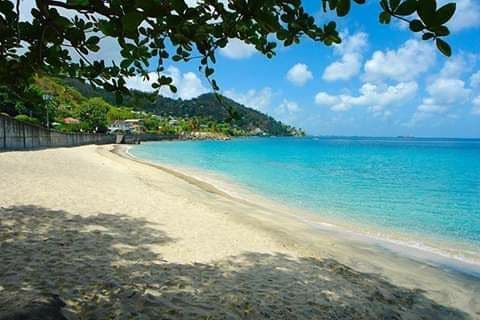A sandy beach extends along the shoreline, with gentle waves lapping at the edge. Lush green hills rise in the background, dotted with colorful homes. The sky is bright blue, filled with scattered fluffy clouds, creating a serene coastal atmosphere.