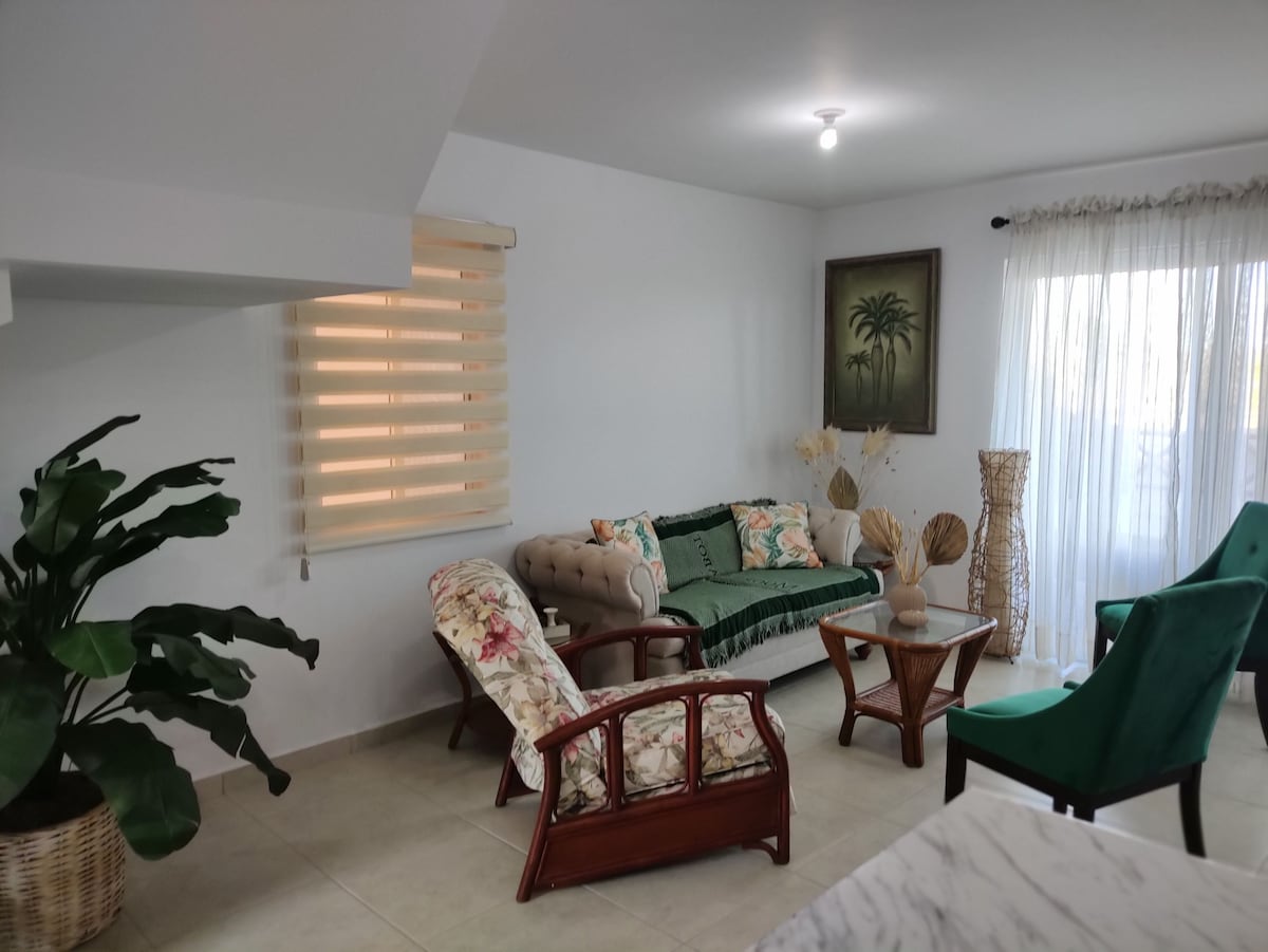 A living area is presented with a combination of seating options, including floral patterned chairs and a tufted sofa. A coffee table sits in the center, surrounded by decorative plants and soft natural light filtering through sheer curtains.
