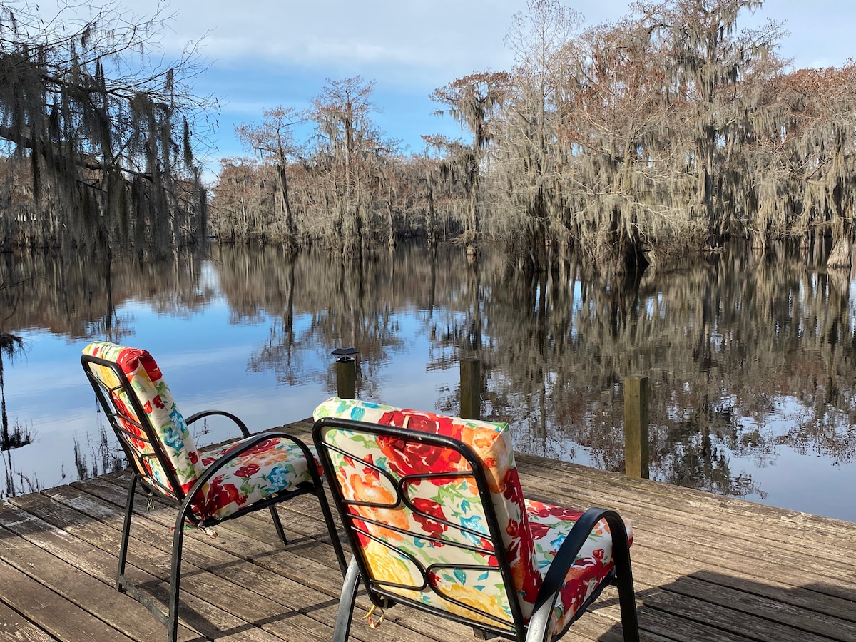 Two colorful lounge chairs are positioned on a wooden deck overlooking a calm lake, surrounded by trees adorned with Spanish moss. The peaceful water reflects the landscape, creating a serene environment for relaxation and contemplation.