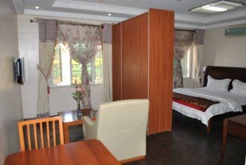 A studio apartment layout is displayed, featuring a spacious sleeping area with a large bed and a partition providing privacy. Natural light enters through two large windows, framed by patterned curtains. A small dining table and chair are positioned beside the bed.