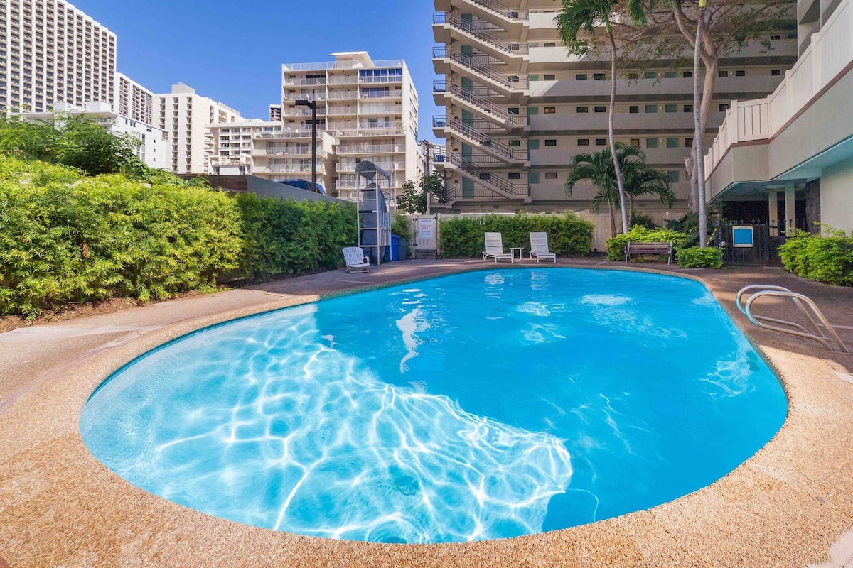 A sunny pool area is depicted, featuring a serene oval-shaped swimming pool with clear blue water. Surrounding the pool, there are lounge chairs positioned for relaxation, framed by lush greenery and tall buildings in the background.