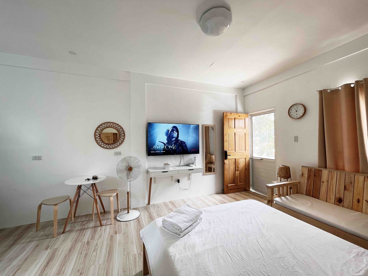 A well-lit studio space features a queen-sized bed, a flat-screen TV mounted on the wall, and a small dining table with two chairs. Natural light fills the room through a window with beige curtains, and wooden accents add warmth to the minimalist design.