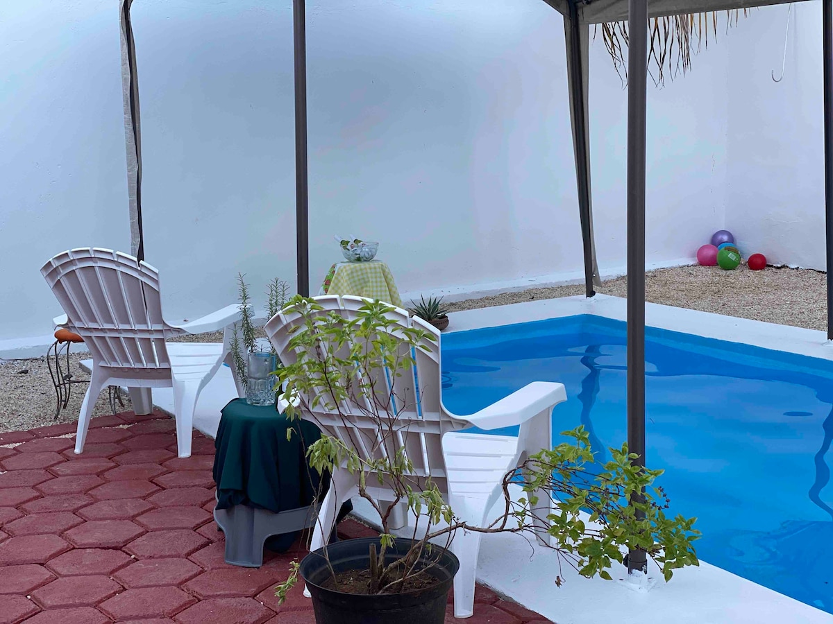A private outdoor area features a small pool surrounded by light-colored walls. Two white chairs are positioned under a gazebo, with a potted plant nearby. A small table is set with a cloth, and colorful balls are visible in the background.