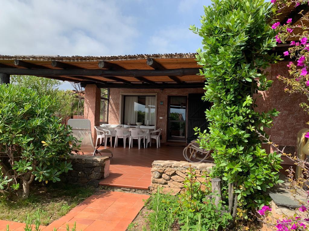 An outdoor patio is featured, furnished with a large dining table surrounded by white chairs. Lush greenery and vibrant flowers frame the space, while a sturdy overhang provides shade. The entrance to the villa is accessible from the patio.