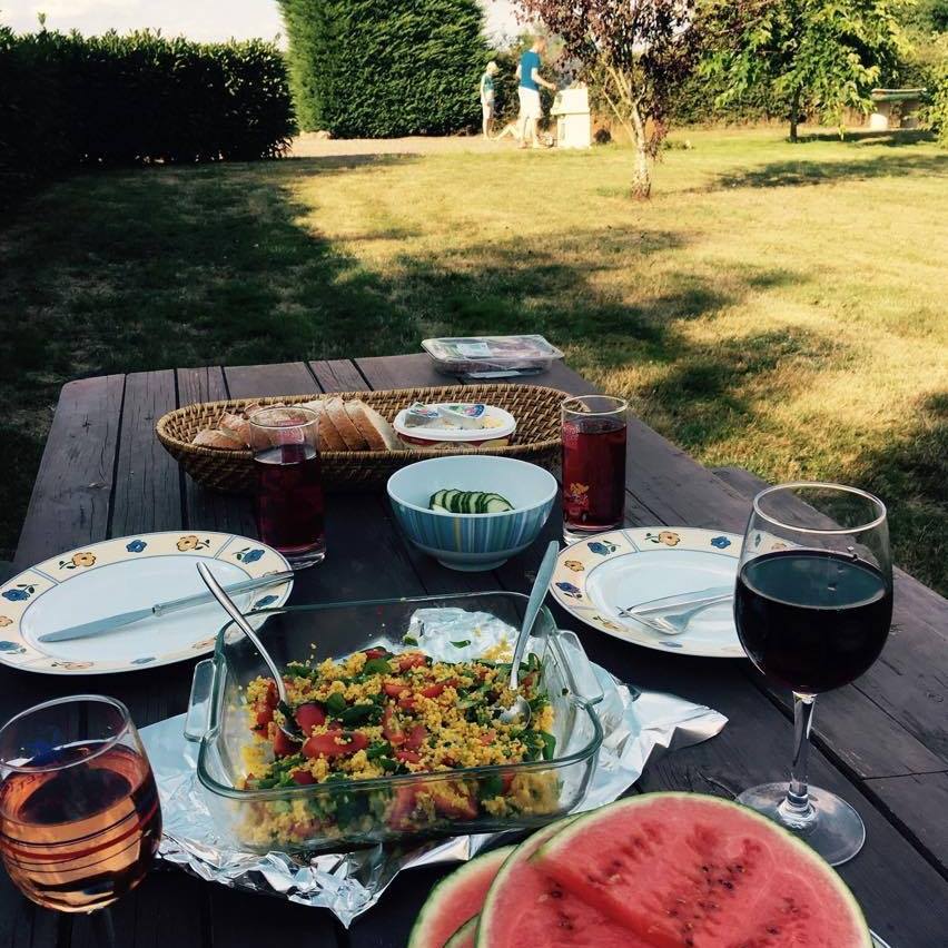 A wooden table is set outdoors with plates, glasses, and a glass dish containing a colorful salad. Watermelon slices are displayed alongside the table. In the background, two individuals are seen walking through the lush green grass, with trees framing the scene.