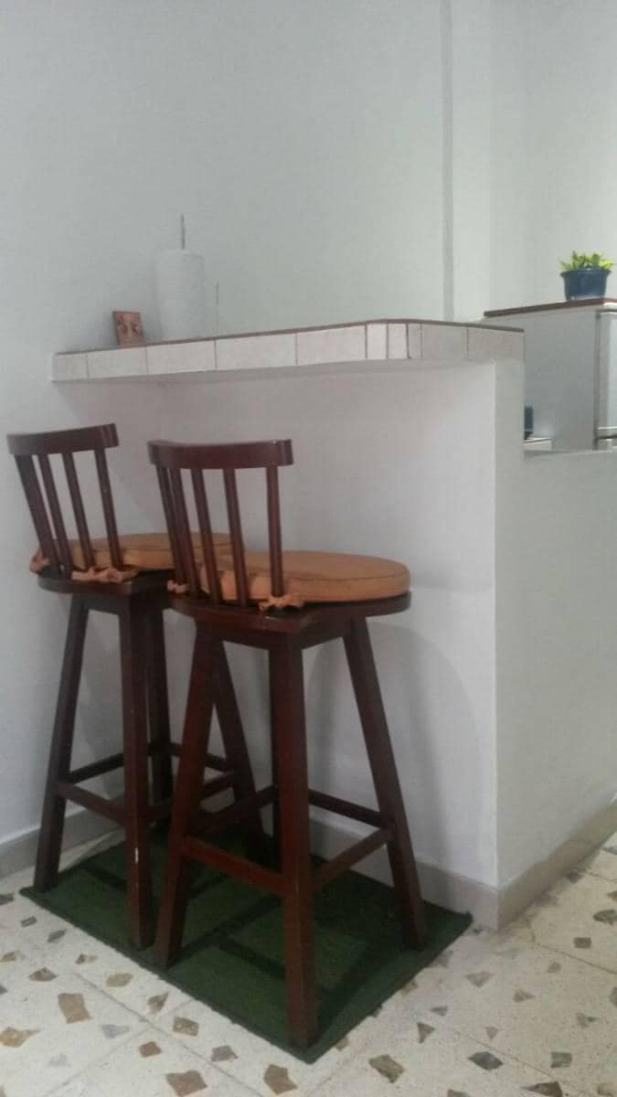 Two wooden bar stools with cushioned seats are positioned at a small counter. The stools feature a simple design, complementing the minimalist decor of the space. A small potted plant is visible on the counter, adding a touch of greenery to the area.