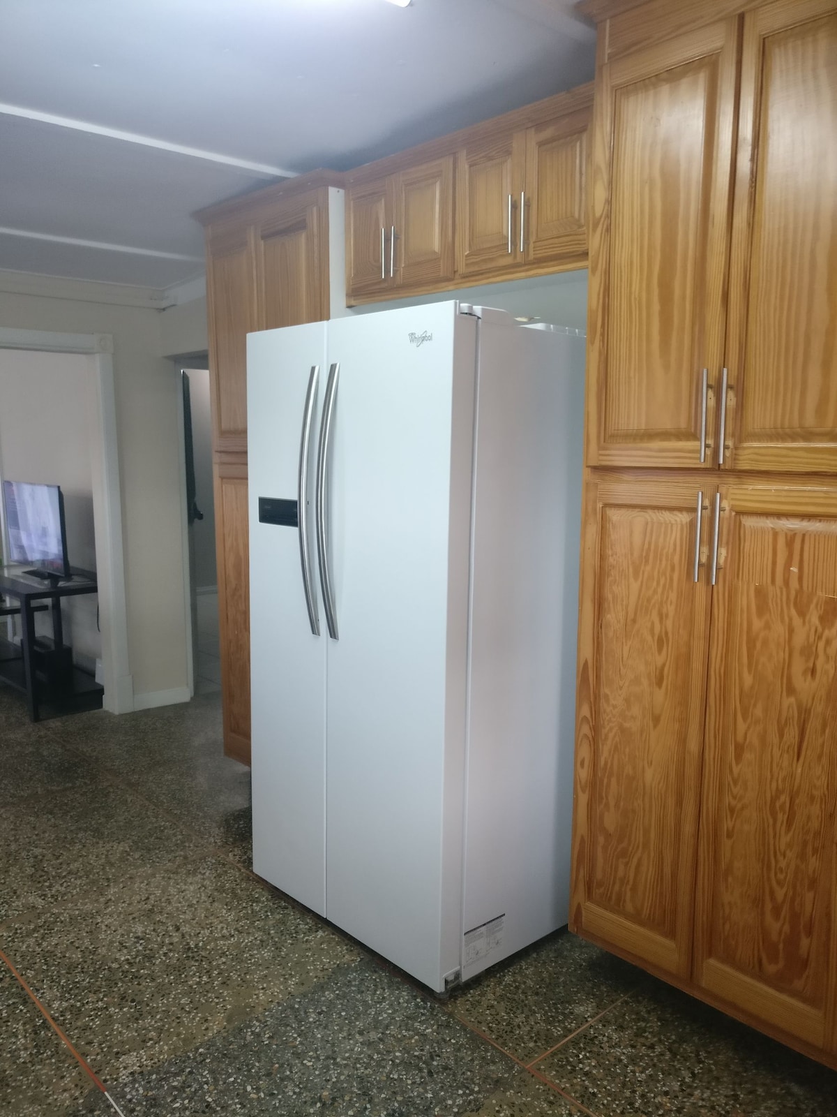 A spacious kitchen area features a sleek white refrigerator with double doors, positioned next to tall wooden cabinets. The cabinets exhibit a warm wood finish, providing ample storage space. The floor is covered in a polished stone surface, enhancing the kitchen's clean and functional look.