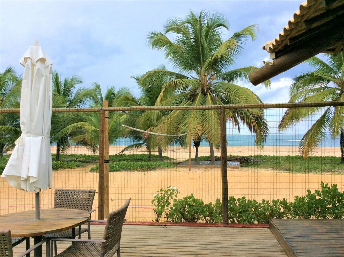 A view of the beach and ocean is framed by swaying palm trees. A wooden deck with outdoor seating is visible in the foreground, featuring a table surrounded by chairs and a white umbrella for shade. The beach and water create a serene backdrop.