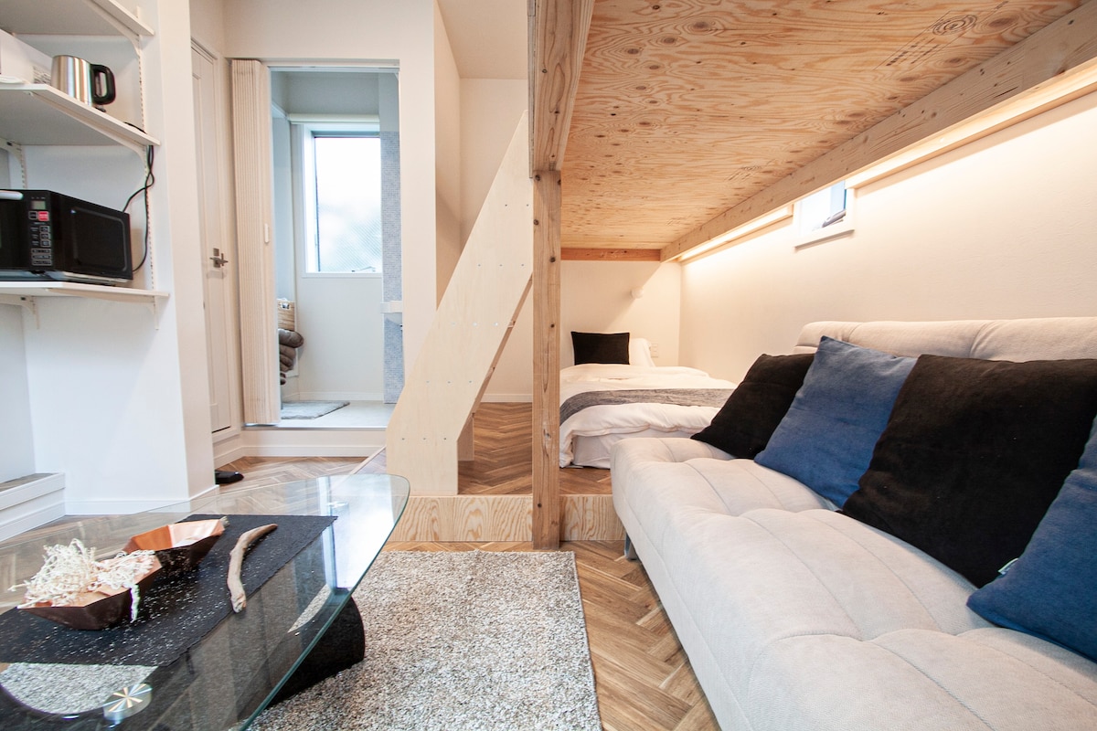 An open-plan layout reveals a cozy sleeping area elevated atop a wooden platform, featuring a softly made bed with a white duvet. A modern sofa in light grey, accented with dark cushions, sits on a textured rug, while a sleek glass table displays decorative items.