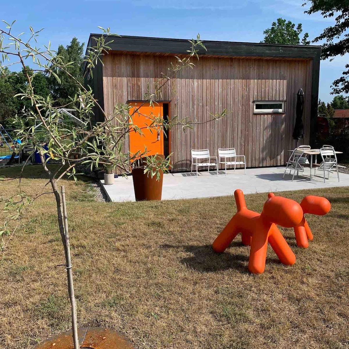 A modern wooden structure is set against a green landscape, featuring a vibrant orange door. A playful orange sculpture shaped like a dog is positioned in the yard, along with two white chairs and a round planter. The area is well-lit with natural sunlight.