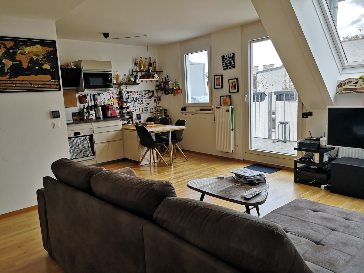 A modern living area features a comfortable sofa facing a coffee table and a sleek entertainment system. A dining area with stylish chairs is situated near the kitchenette, while large windows provide ample natural light and a view of the terrace beyond.