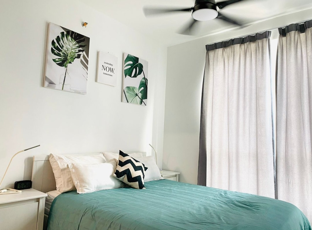 The bedroom features a spacious bed adorned with a teal duvet and decorative cushions. Two artworks featuring botanical prints are displayed above the bed. A window is partially covered by textured gray curtains, and a ceiling fan provides effective air circulation.
