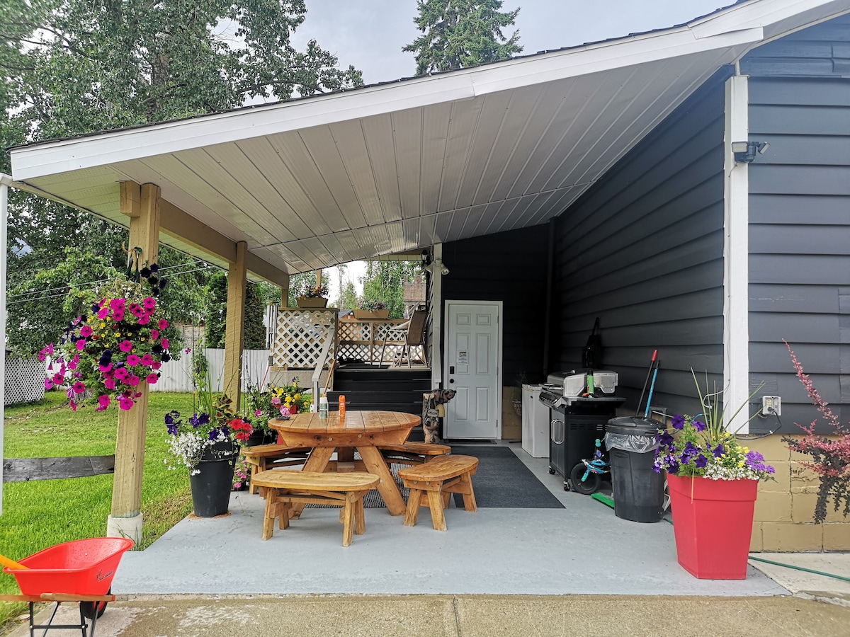 An outdoor covered patio is presented with a wooden picnic table surrounded by benches. Colorful flower pots and hanging baskets add vibrancy, while a barbecue grill and storage area are visible nearby. The space offers a relaxing setting for outdoor gatherings.