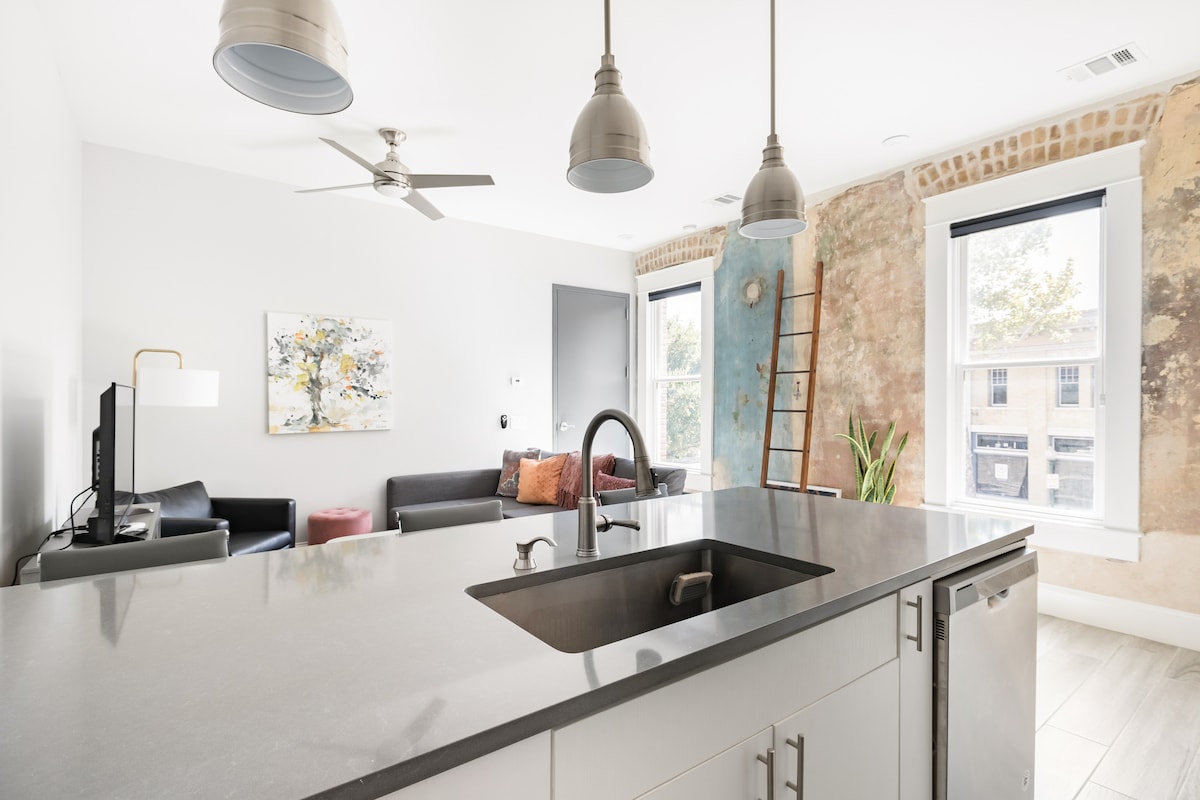 A modern kitchen features sleek countertops and a stainless steel sink. In the background, a living area displays a minimalist design with dark sofas, a vibrant art piece, and large windows showcasing natural light, while the exposed brick wall adds character to the space.