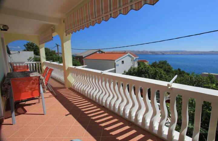 A spacious balcony is highlighted with a dining table surrounded by four orange chairs. The railing offers a view of the clear blue sea and distant hills under bright sunlight, creating a welcoming area for relaxation.