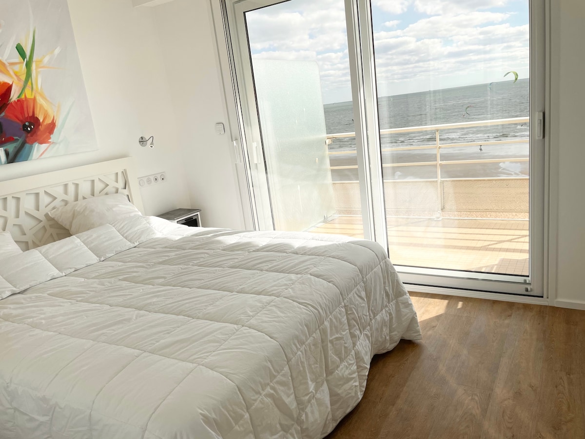 A bright bedroom features a spacious bed covered with a white quilt. A large sliding door leads to a balcony, offering an unobstructed view of the ocean. Natural light fills the room, and a floral artwork hangs on the wall, adding a touch of color.