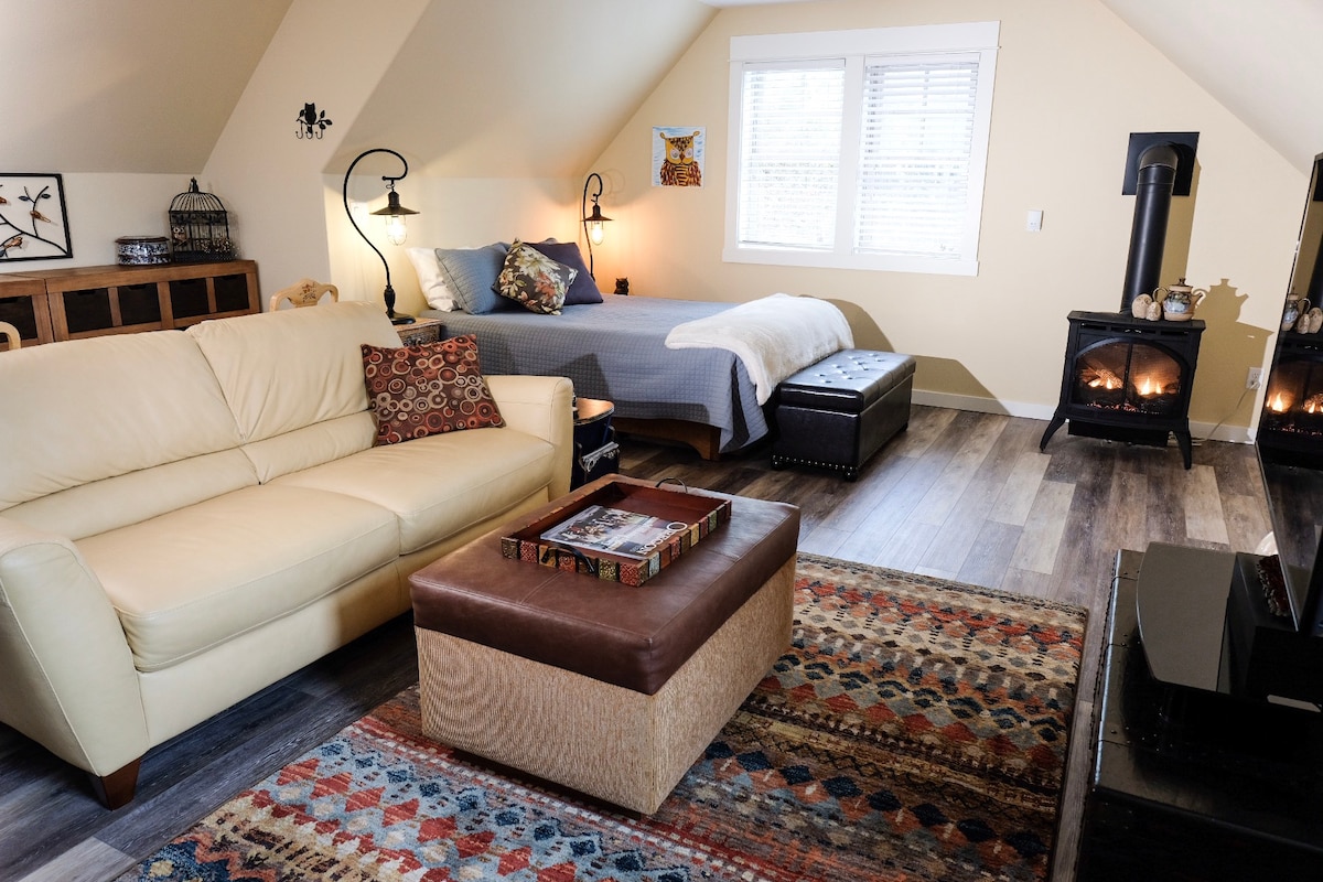 A spacious studio apartment is presented with a comfortable cream-colored couch and a patterned area rug. A queen-sized bed is visible in the background, accompanied by a cozy wood stove. Natural light enters through a window, enhancing the neutral-toned walls.