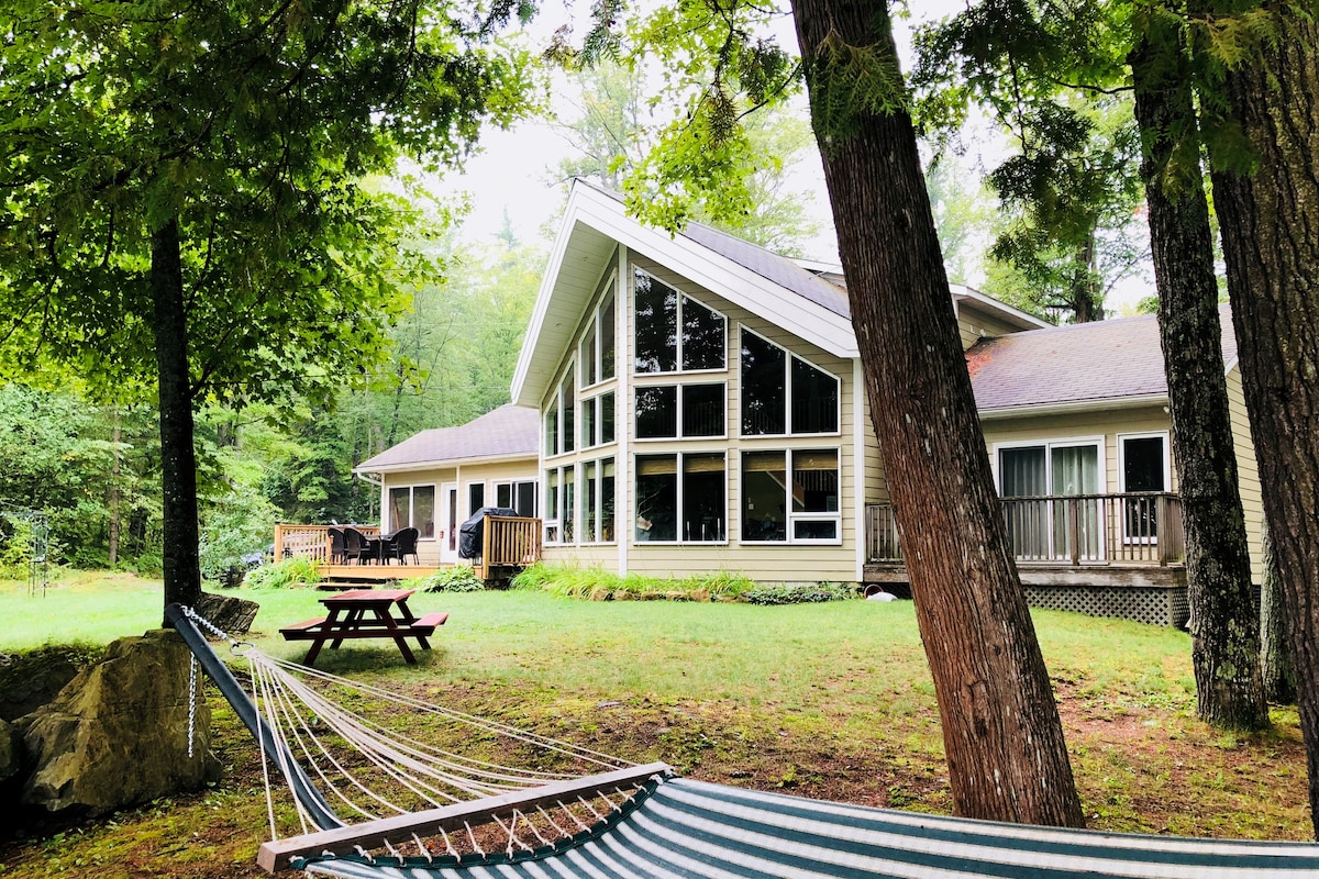 The image features a spacious cottage surrounded by trees, showcasing large windows and a peaked roof design. A hammock is positioned in the foreground, with picnic tables and a deck visible in the background, providing a relaxing outdoor space near the lake.