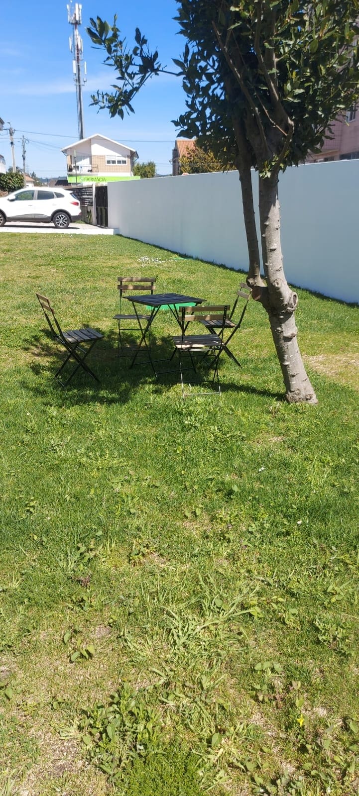 An outdoor space features a grassy area with a small table surrounded by four chairs. A tree provides partial shade, creating a simple setting for relaxation or dining. The background includes a white fence and glimpses of nearby vehicles.
