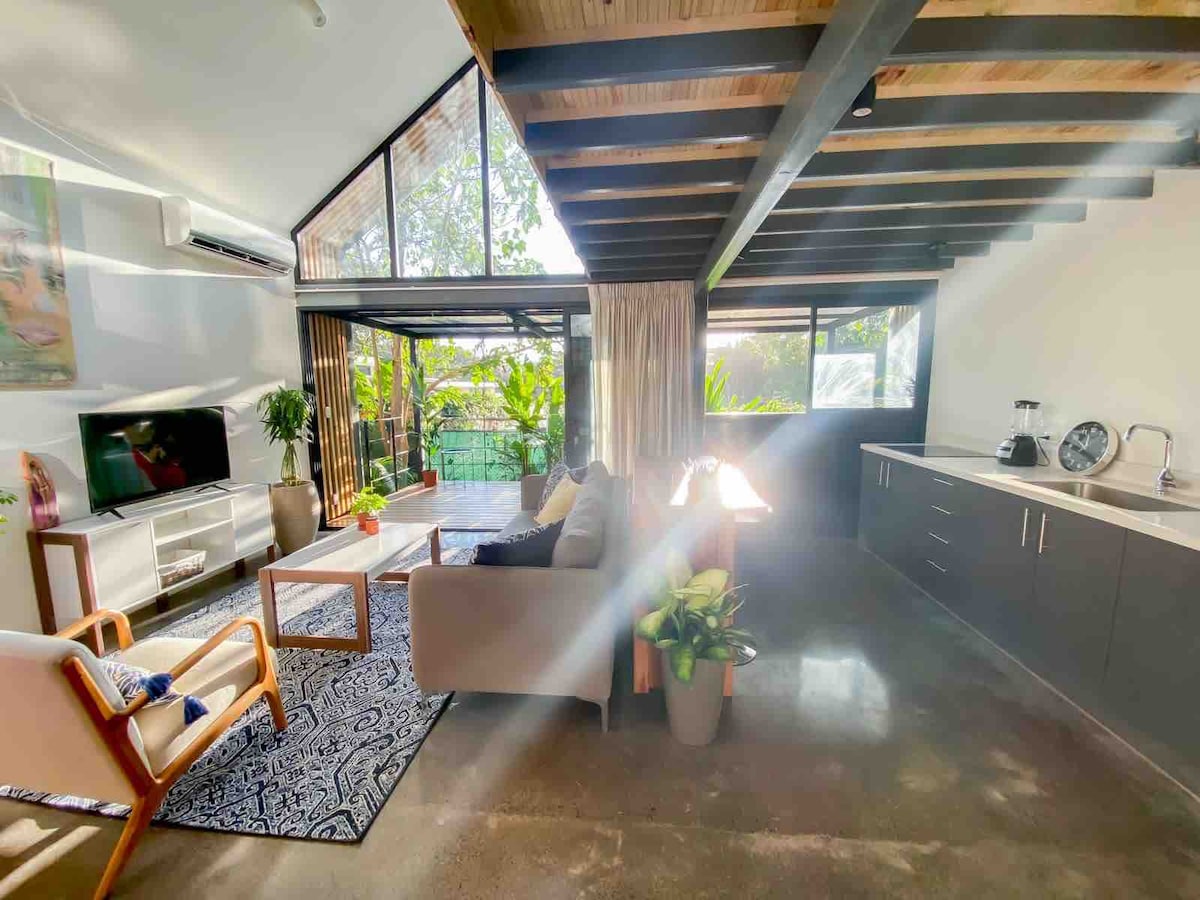 A bright living area is presented with large glass doors opening to a lush outdoor view. Sleek kitchen cabinetry complements the space, while a cozy sofa and wooden coffee table invite relaxation. Natural light streams through, enhancing the modern wood-beamed ceiling.