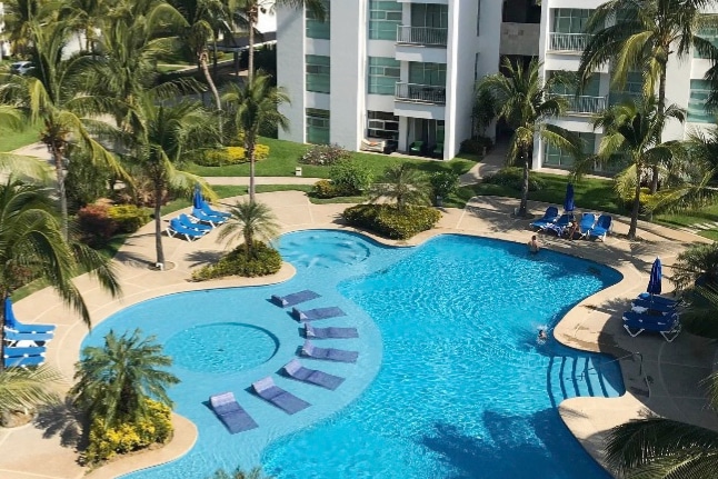 A large swimming pool features a circular area with a shallow section and multiple loungers arranged around it. Lush palm trees frame the pool area, complemented by landscaped gardens with vibrant flowers. The surrounding building has multiple balconies overlooking the pool.
