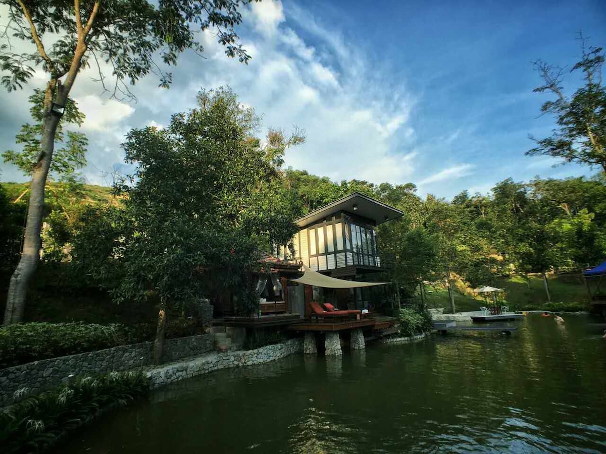 A villa is situated on stilts over a serene pond, surrounded by lush greenery. Large windows provide views of the natural environment, while a shaded seating area with chairs offers a place to relax by the water. The sky showcases a mix of clouds and blue tones.