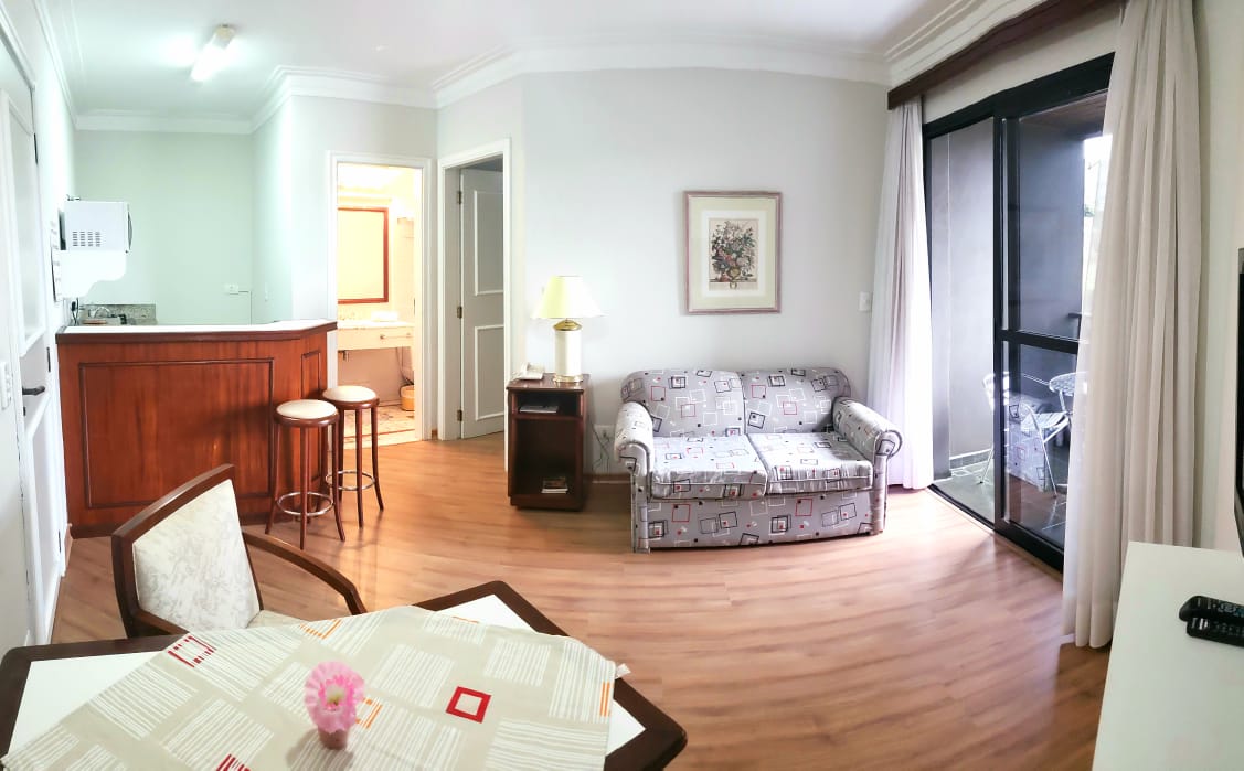 The living area features a patterned sofa and a dining table with light-colored tabletop. A bar area is visible in the background, with two stools. Natural light filters in through large glass doors that lead to a private balcony, enhancing the spacious feel.