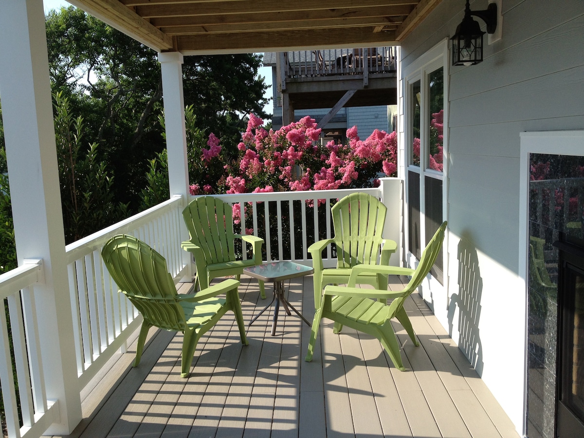 A covered porch is highlighted by four green Adirondack chairs surrounding a small table. Lush pink flowering bushes provide a vibrant backdrop, while the wooden ceiling and flooring offer a natural touch to the inviting outdoor space.