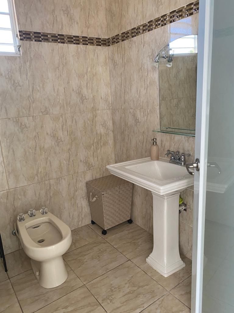 A bathroom is seen featuring beige tiled walls and floor. A single pedestal sink is positioned next to a wall-mounted mirror. A bidet is located adjacent to the toilet, providing essential amenities in a compact layout.