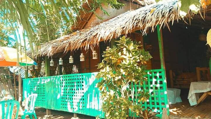 A traditional Bahay Kubo features a thatched roof and wooden structure, surrounded by lush greenery. A vibrant teal railing surrounds the porch, where decorative lanterns hang. Sunlight filters through the leaves, creating an inviting atmosphere in this outdoor space.