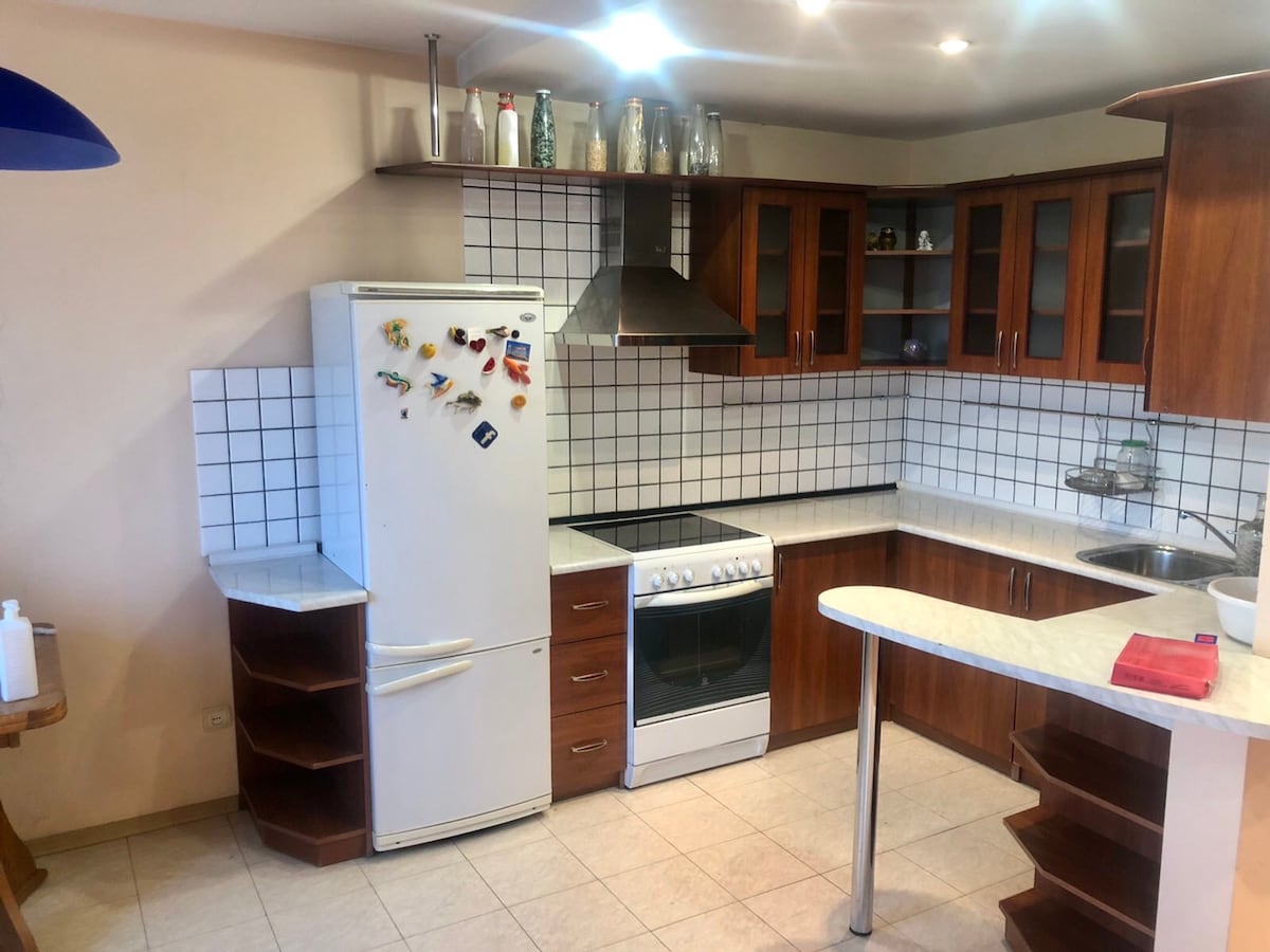 A kitchen space is presented, featuring wooden cabinetry and a white tiled backsplash. A refrigerator stands against one wall, beside a freestanding oven with a stovetop. Shelves display various kitchen items, while a sink is positioned next to the counter, offering ample workspace.