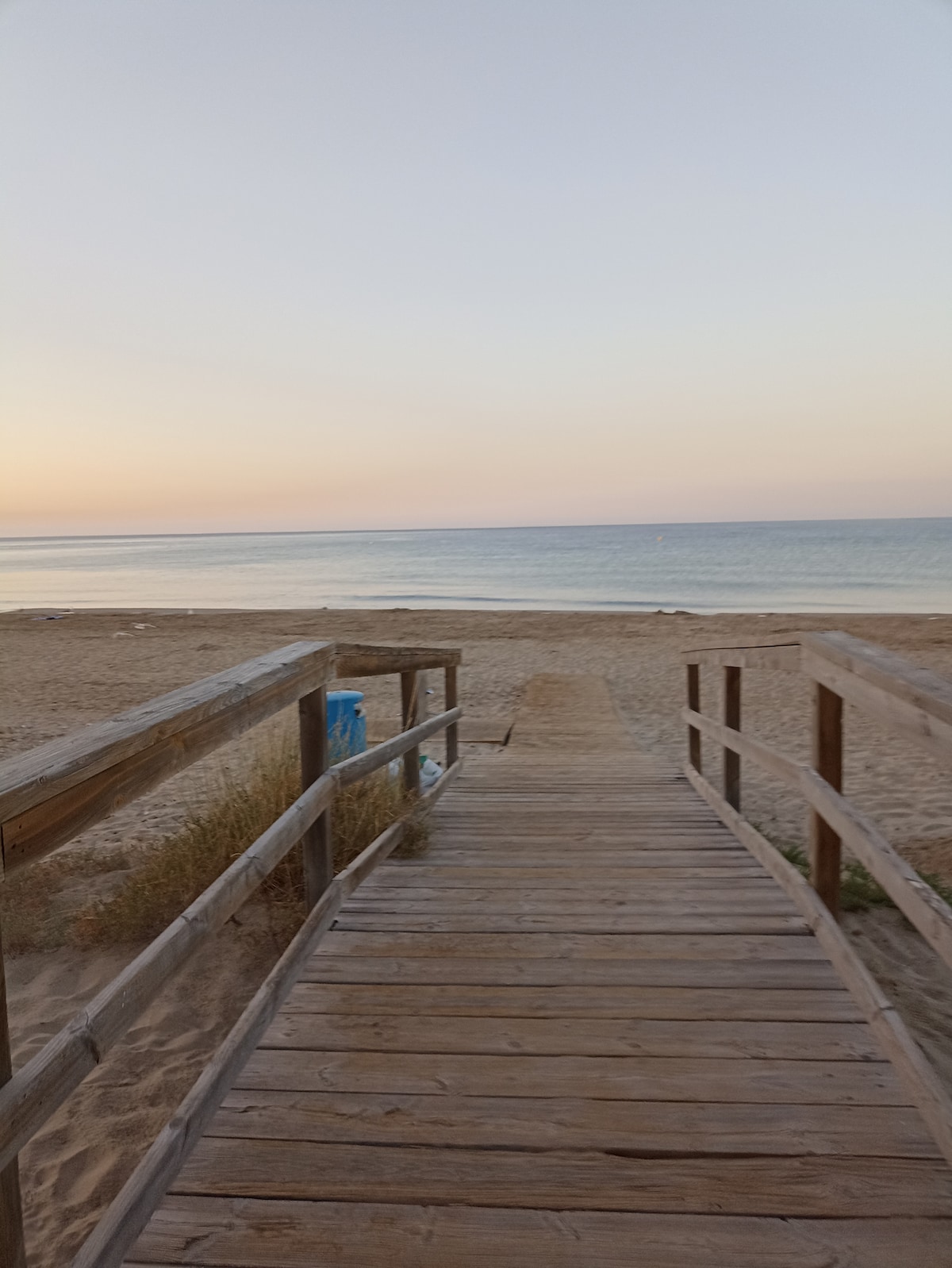 A wooden path extends towards a tranquil beach, leading to calm waters with soft reflections of pastel hues in the sky. The sandy shore is visible on both sides, with gentle waves lapping at the edge, creating a serene atmosphere.