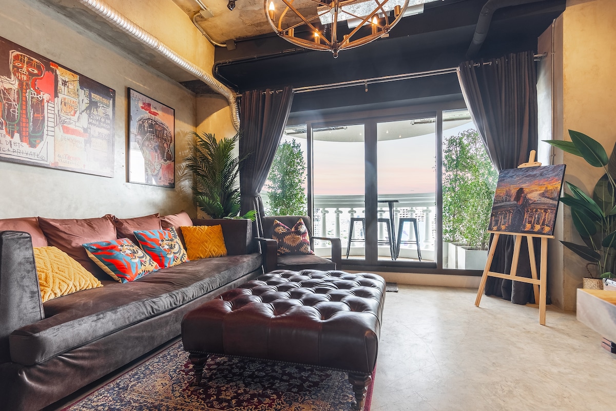 A spacious living room features a large, comfortable sofa adorned with colorful throw pillows. An elegant chandelier hangs from the ceiling, and sliding glass doors lead to a private balcony with bar seating. Nearby, a decorative easel displays artwork against a backdrop of potted greenery.