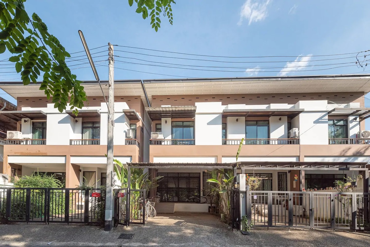 The exterior of the townhouse features a modern design with multiple levels. Large windows allow natural light to fill the interiors. Greenery adorns the front entrance, enhancing the welcoming atmosphere. A black metal fence encloses the property, while air conditioning units are visible on each side.