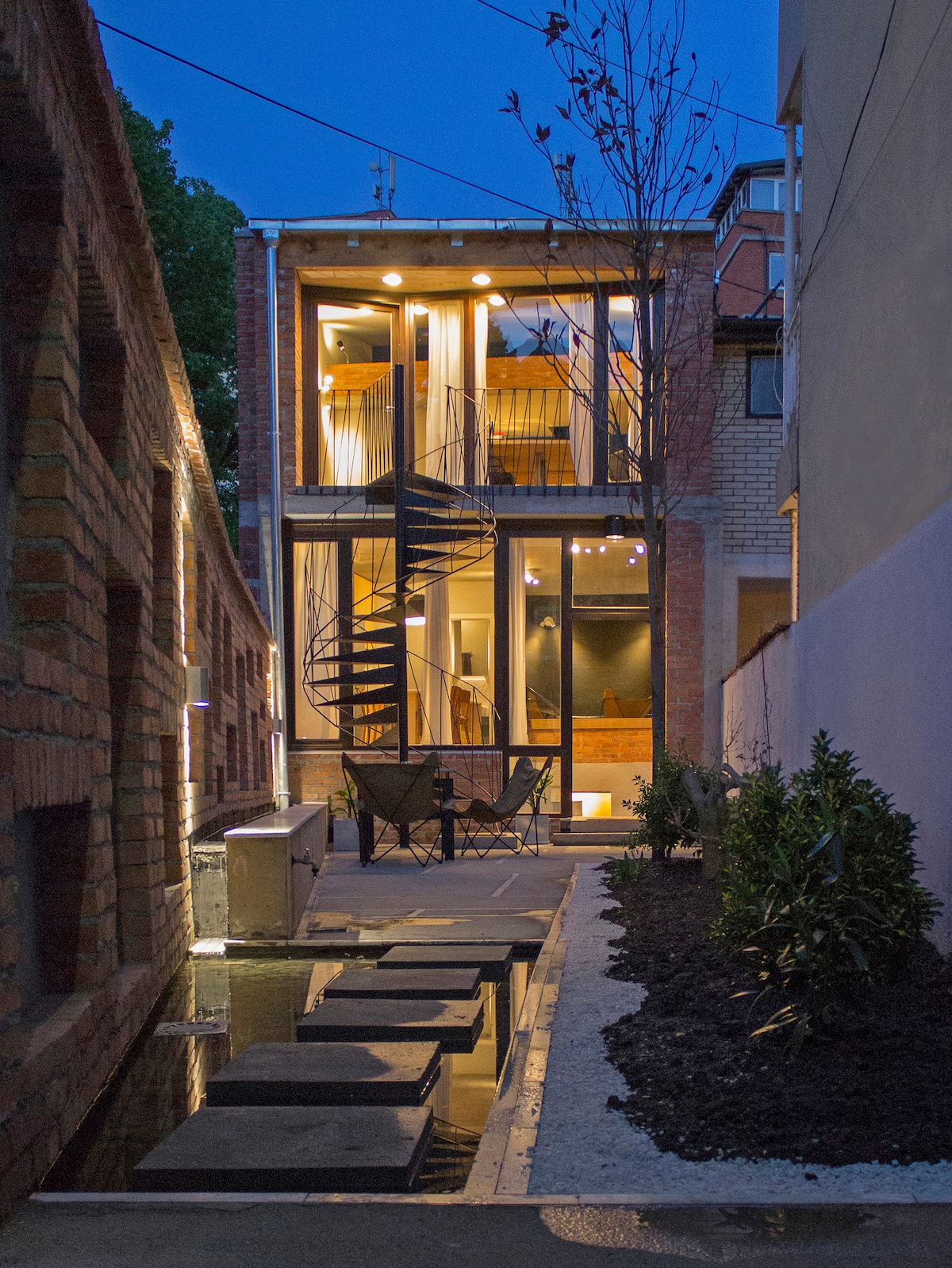 An exterior view of the two-story house is captured at dusk, featuring a spiral staircase leading to a balcony. Large windows illuminate the interior, while a narrow walkway with water features reflects the structure. Surrounding greenery adds a natural touch to the urban setting.