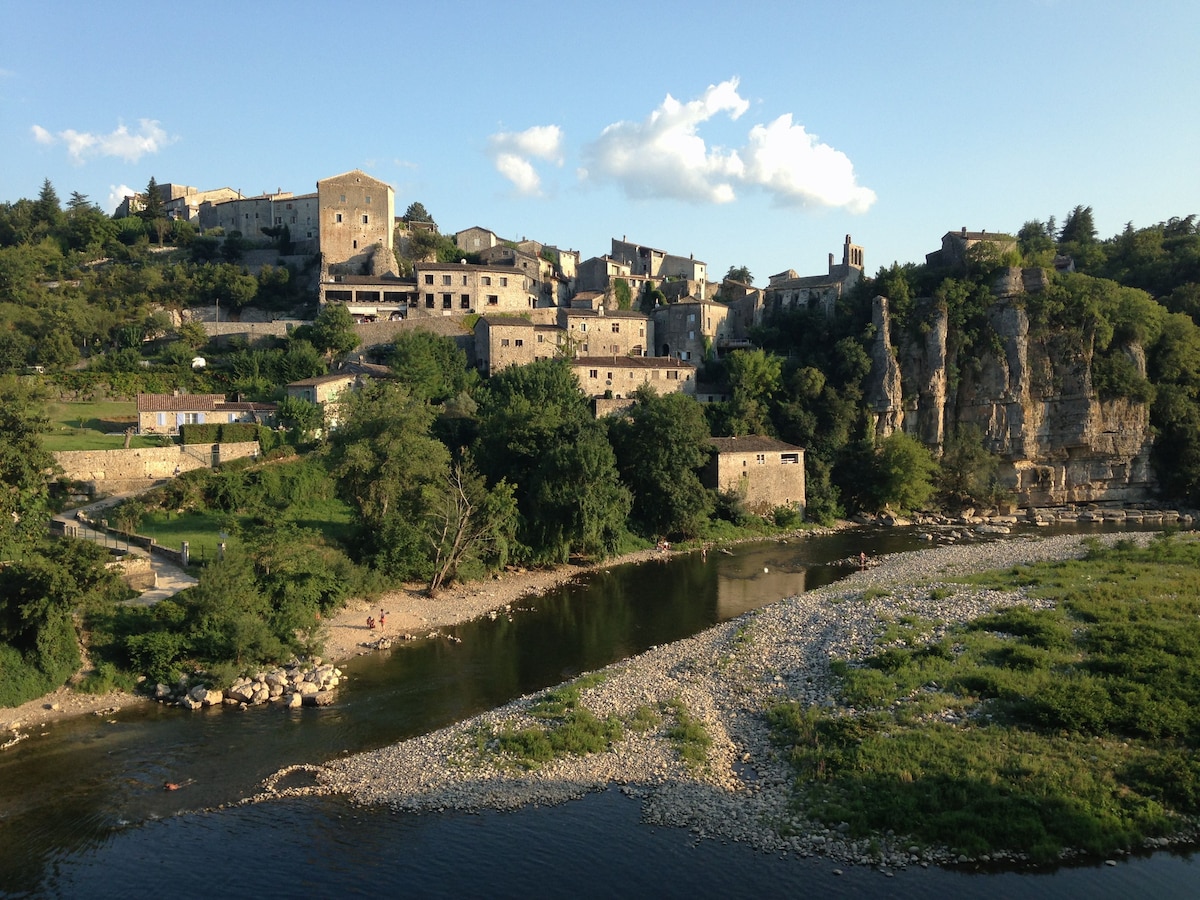 A historic village is nestled on a hillside, with stone buildings reflecting sunlight. The surrounding landscape features a river flowing gently at the base, bordered by pebbles and lush greenery. Cliffs rise prominently beside the village, accentuating the natural beauty of the area.