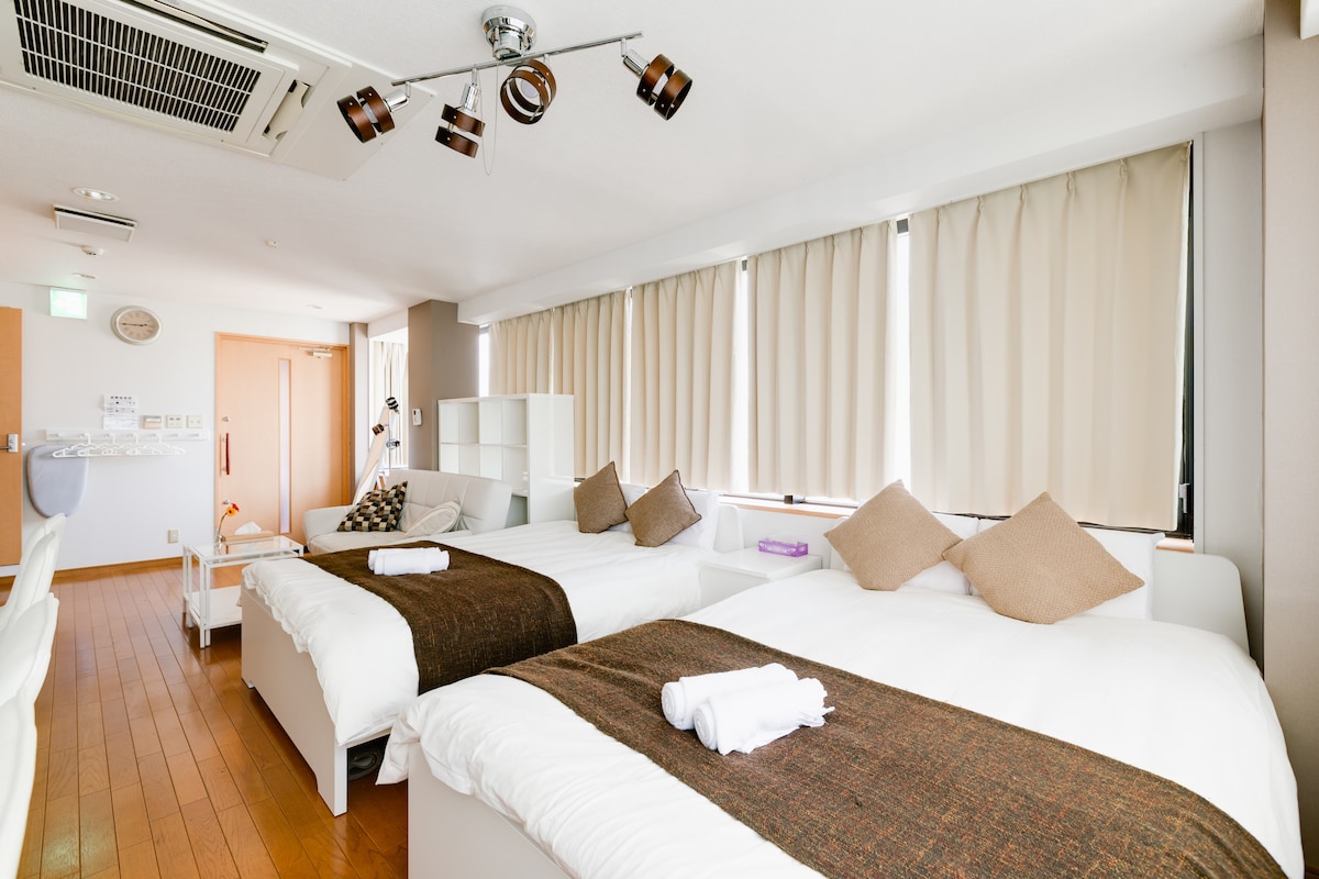A room is furnished with two spacious beds, each adorned with neutral-toned blankets and decorative pillows. Large windows with light-colored curtains allow natural light to fill the space. A comfortable seating area is visible in the background, along with minimalist decor.