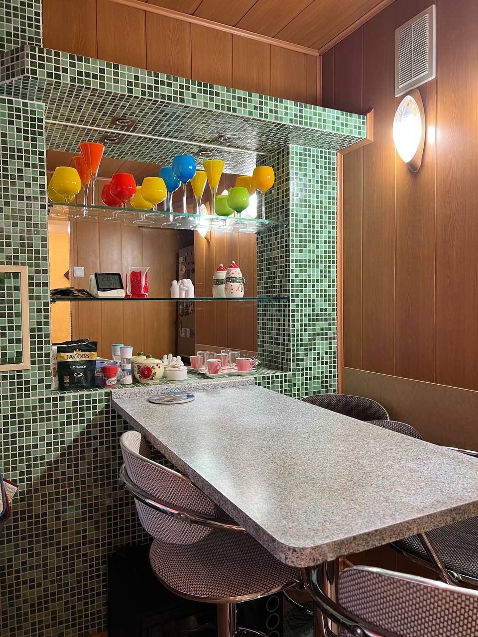 A modern bar area features a long rectangular table with high stools. Colorful glassware hangs above in a playful display, while tiled walls in shades of green provide a vibrant backdrop. The well-lit space offers a welcoming atmosphere for gatherings.