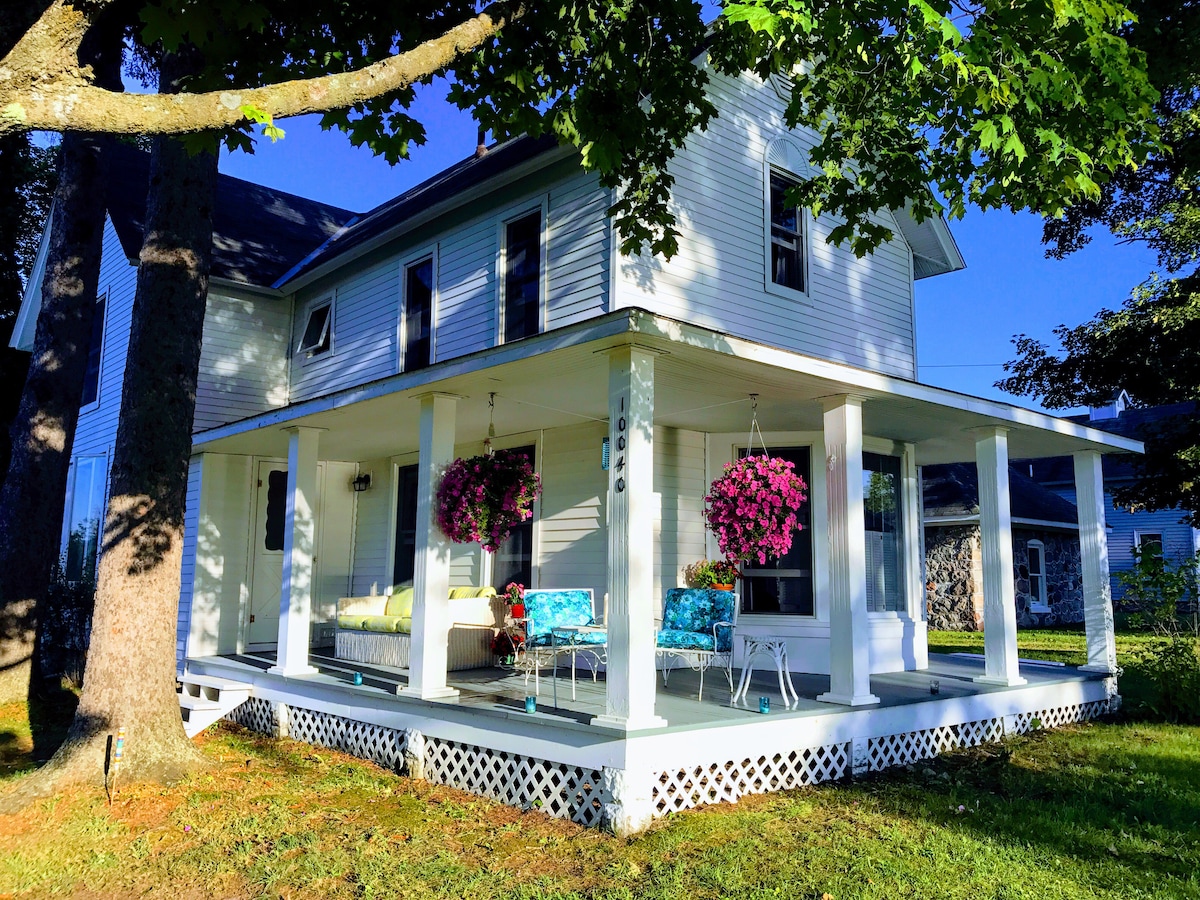 A vintage farmhouse is featured with a wrap-around porch, adorned with hanging flower baskets. Bright seating arrangements are placed on the porch, allowing for relaxation while enjoying the surrounding greenery. Large windows reflect natural light, creating an inviting outdoor space.